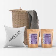 pack of SoluCLEAN Non Bio Laundry Detergent sachets and SoluCLEAN Bio Laundry Detergent sachets with purple labels, a small white cushion with the soluCLEAN logo is next to them, and behind them all is a light brown wicker basket with a towel hanging out 